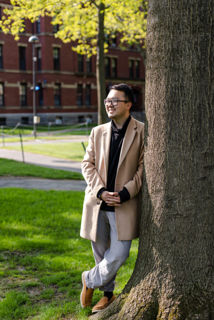 Charles Hua, a Harvard undergraduate student in the class of 2023, smiles for a photo while leaning against a tree in Harvard Yard.
