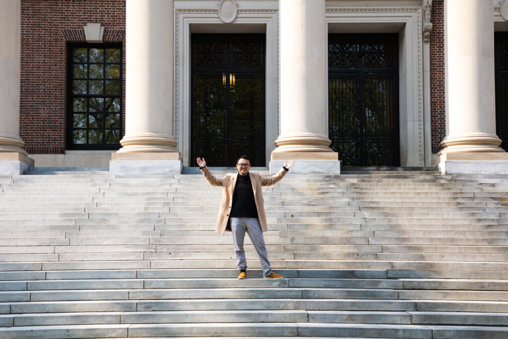 Charles Hua, a Harvard undergraduate student in the class of 2023, lifts his hands in the air while standing on the steps of the Widener Library.