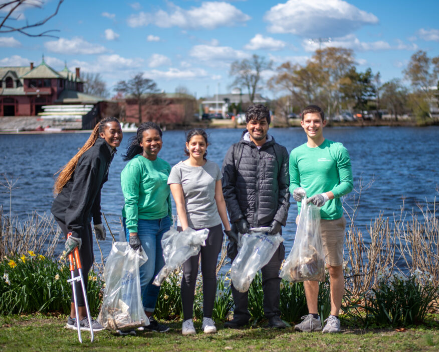 Students pose for a photo near the river holding trash removal tools during the 2022 Charles River Clean-Up event.