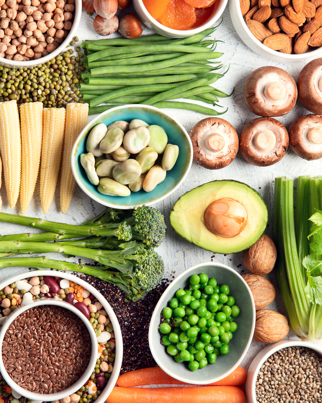 Plant-based foods that are good sources of plant based protein are laid on a table. Image includes avocados, lentils, broccolini, chickpeas, carrots, mushrooms and more.