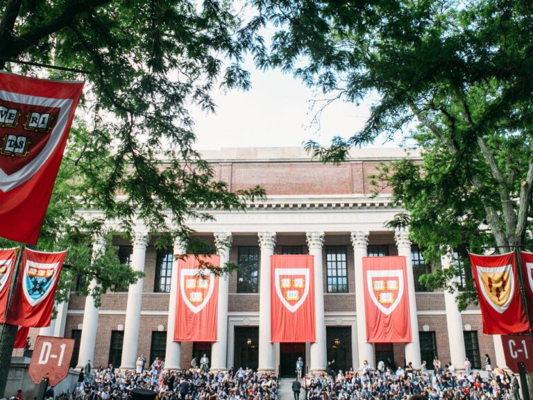 Commencement ceremony photo at Harvard. Several crimson banners with the Harvard shield logo in front can be seen on a building with columns.