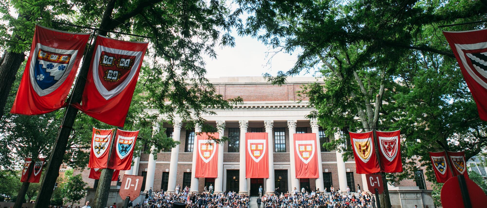 Commencement ceremony photo at Harvard. Several crimson banners with the Harvard shield logo in front can be seen on a building with columns.