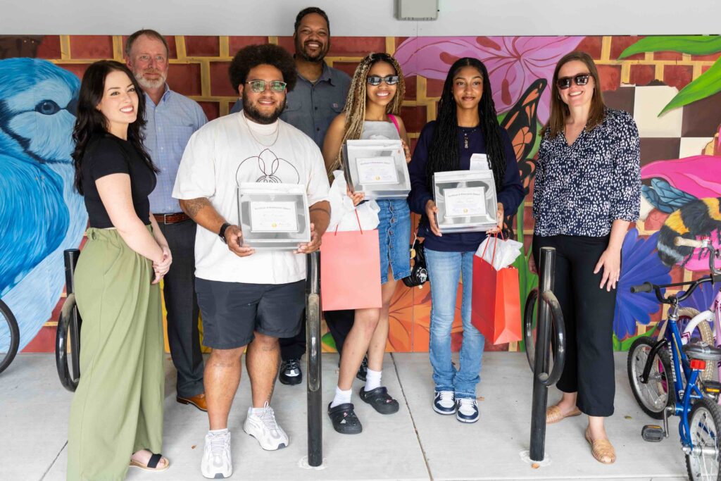 Student artists pose for a photo with Harvard Housing and operational staff. The artists hold a certificate recognizing their work for a mural they painted at a Harvard residential building.