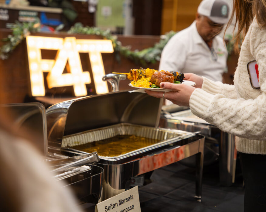 A person in a white sweater is holding a plate of food at a buffet, selecting items from a chafing dish labeled "Seitan Marsala." In the background, a person in a white shirt and hat stands behind the buffet setup. There is a decorative sign made of lightbulbs with the text "F4T" and some greenery displayed. The atmosphere suggests a catered event or food festival.